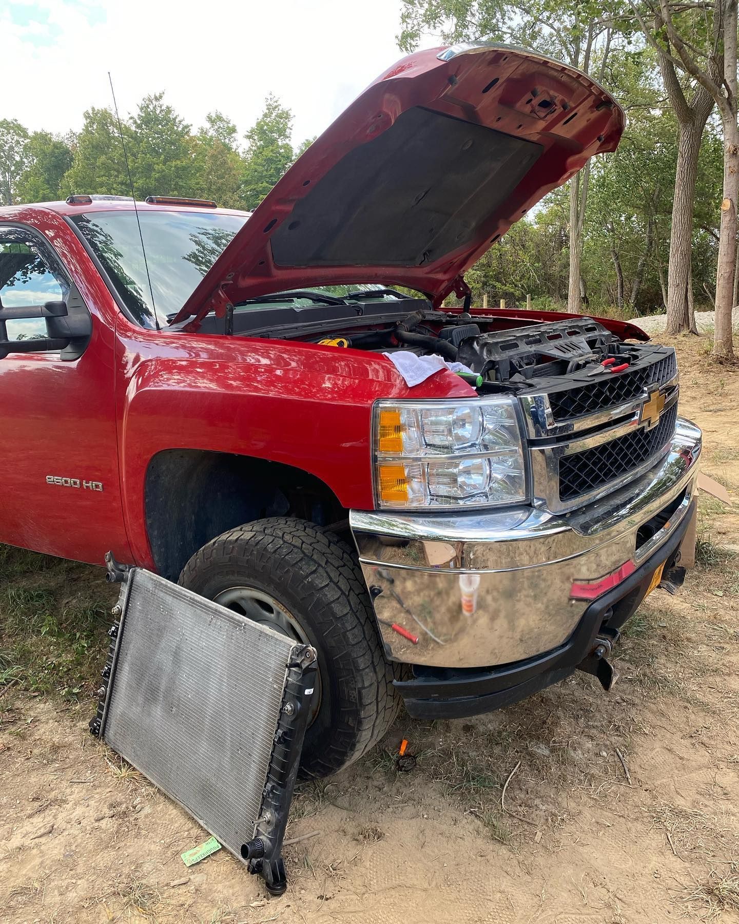 Red pickup truck with hood open and damaged front end, with a detached radiator on the ground in a wooded area