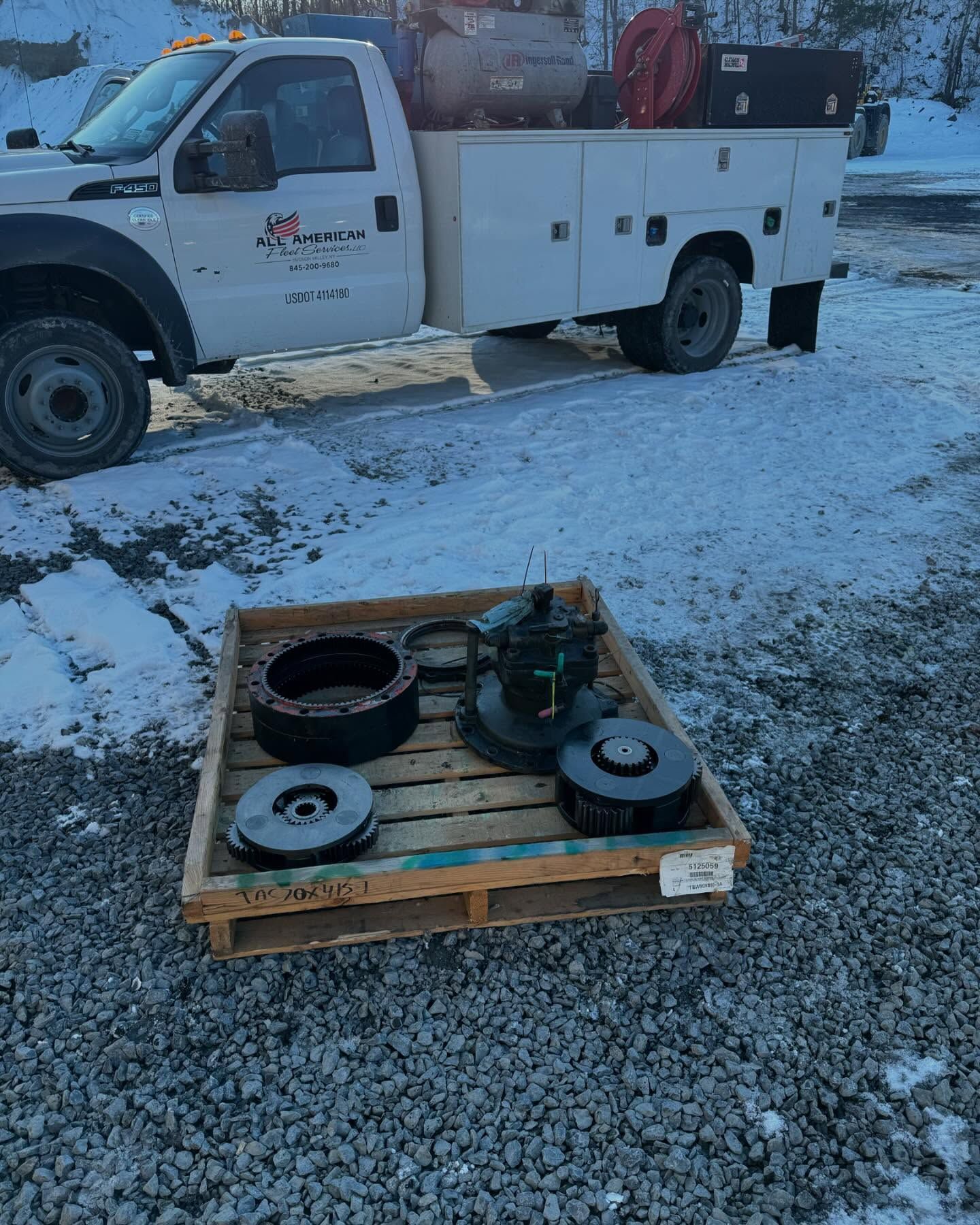 White utility truck beside a pallet with metal parts on a snowy gravel lot