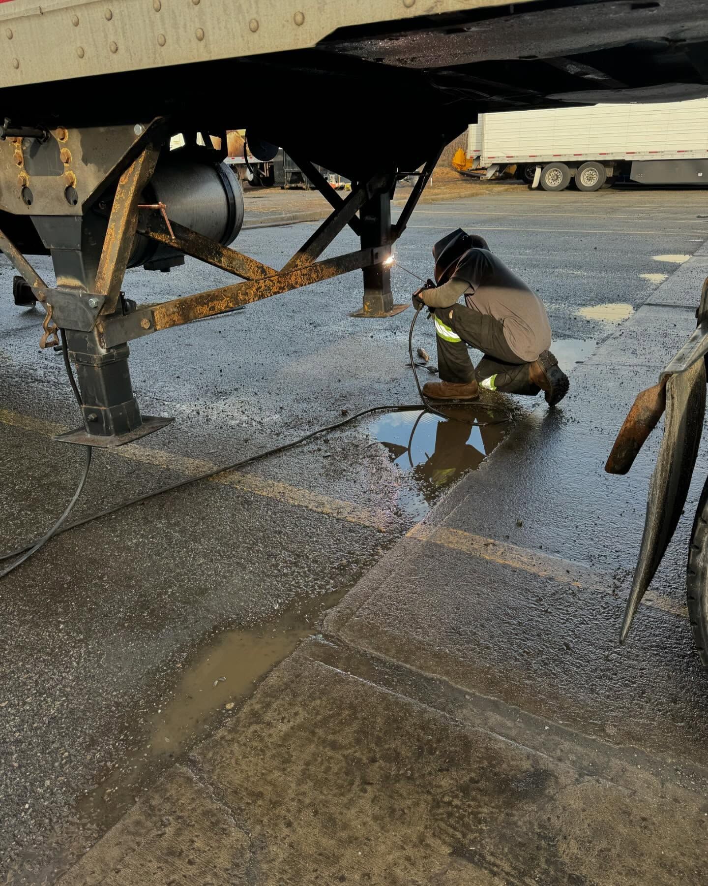 Worker crouching under a trailer, inspecting a wet pavement area with puddles and gravel.