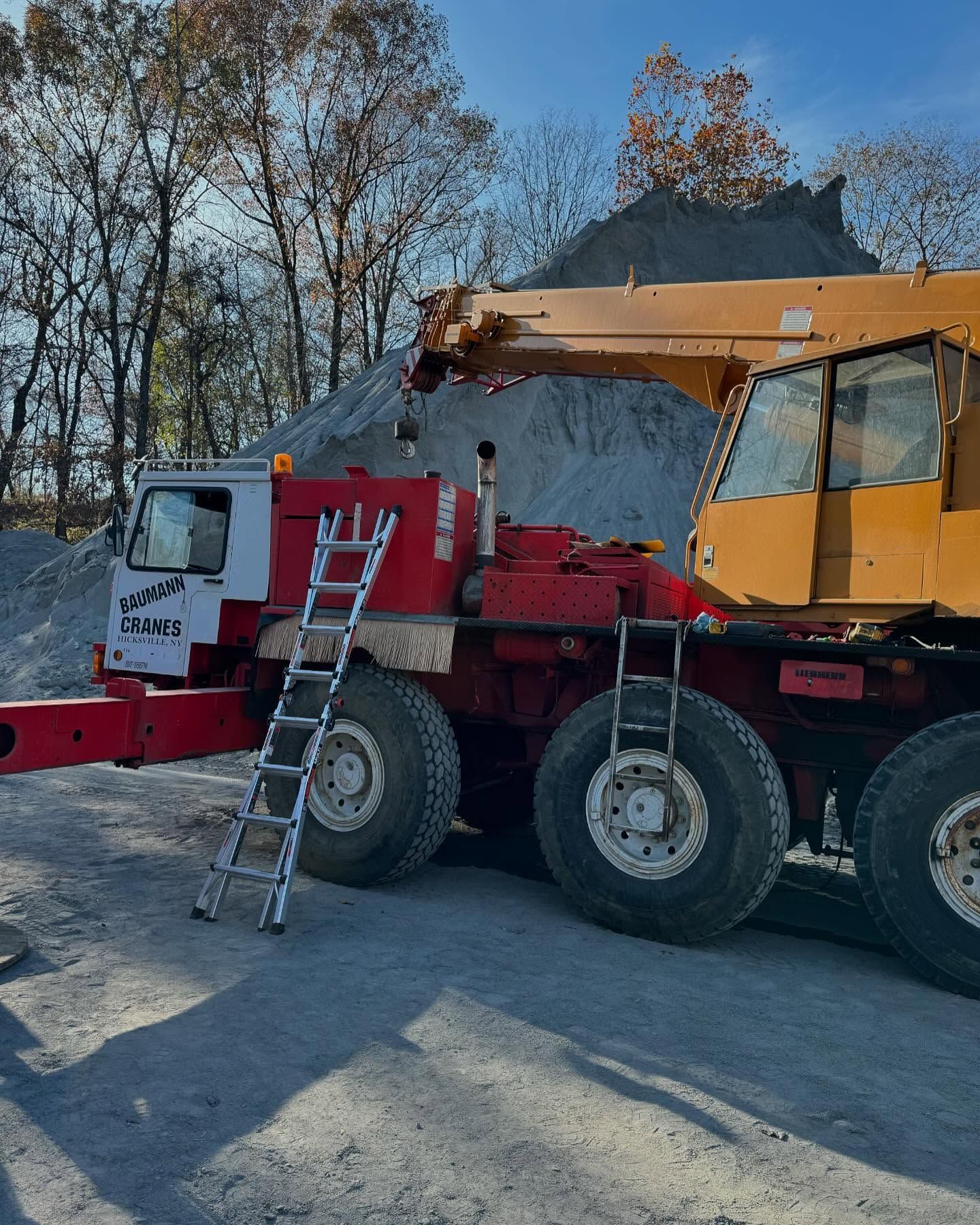 Crane truck loading gravel, with a ladder leaning against its red cab in a snowy worksite