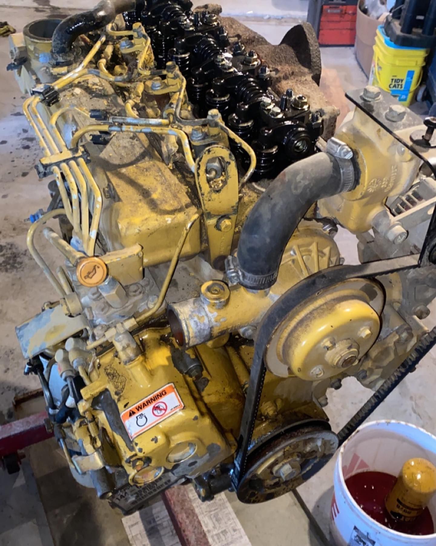 Yellow diesel engine with exposed components and black hoses in a workshop, with a red container nearby