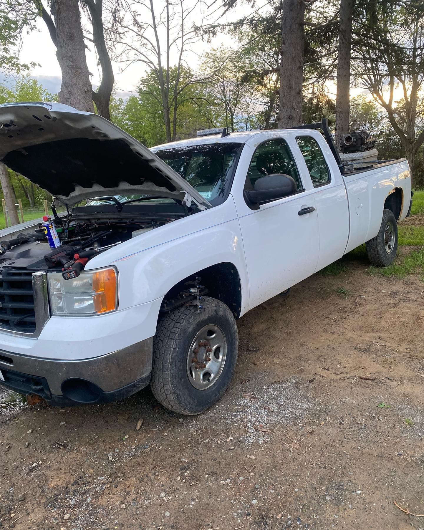 White pickup truck with hood open, parked on a gravel area near trees at dusk