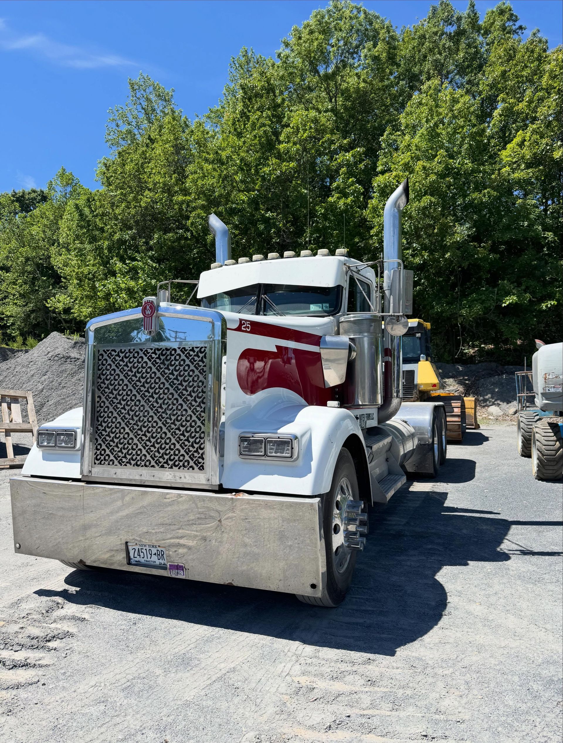 White semi-truck parked on gravel under trees, front view with chrome grille and tall exhaust stacks.