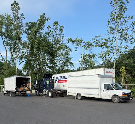 Two white box trucks and a black semi parked in a lot beside tall trees on a sunny day