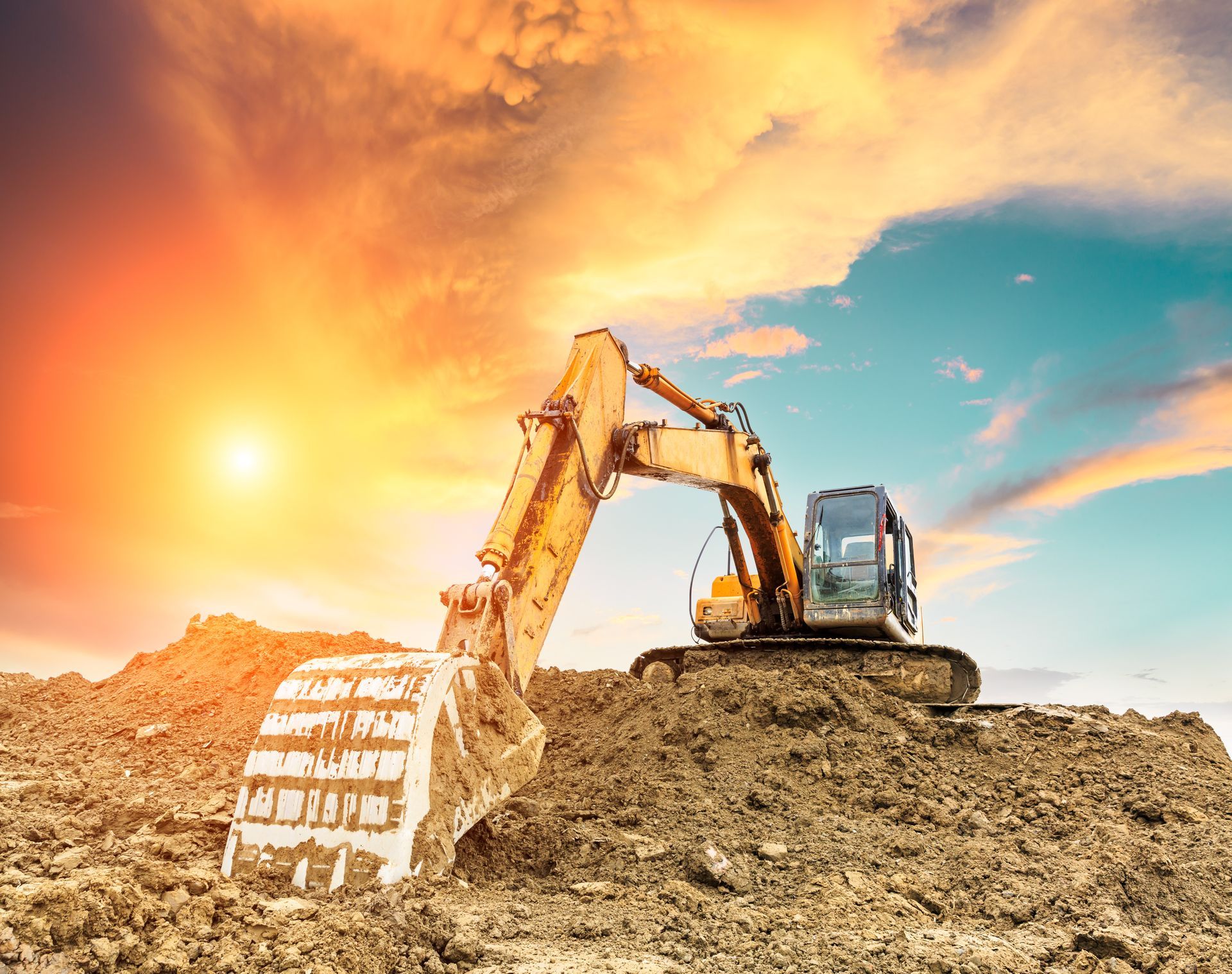 Excavator on a dirt mound at sunset, digging earth under a bright orange sky