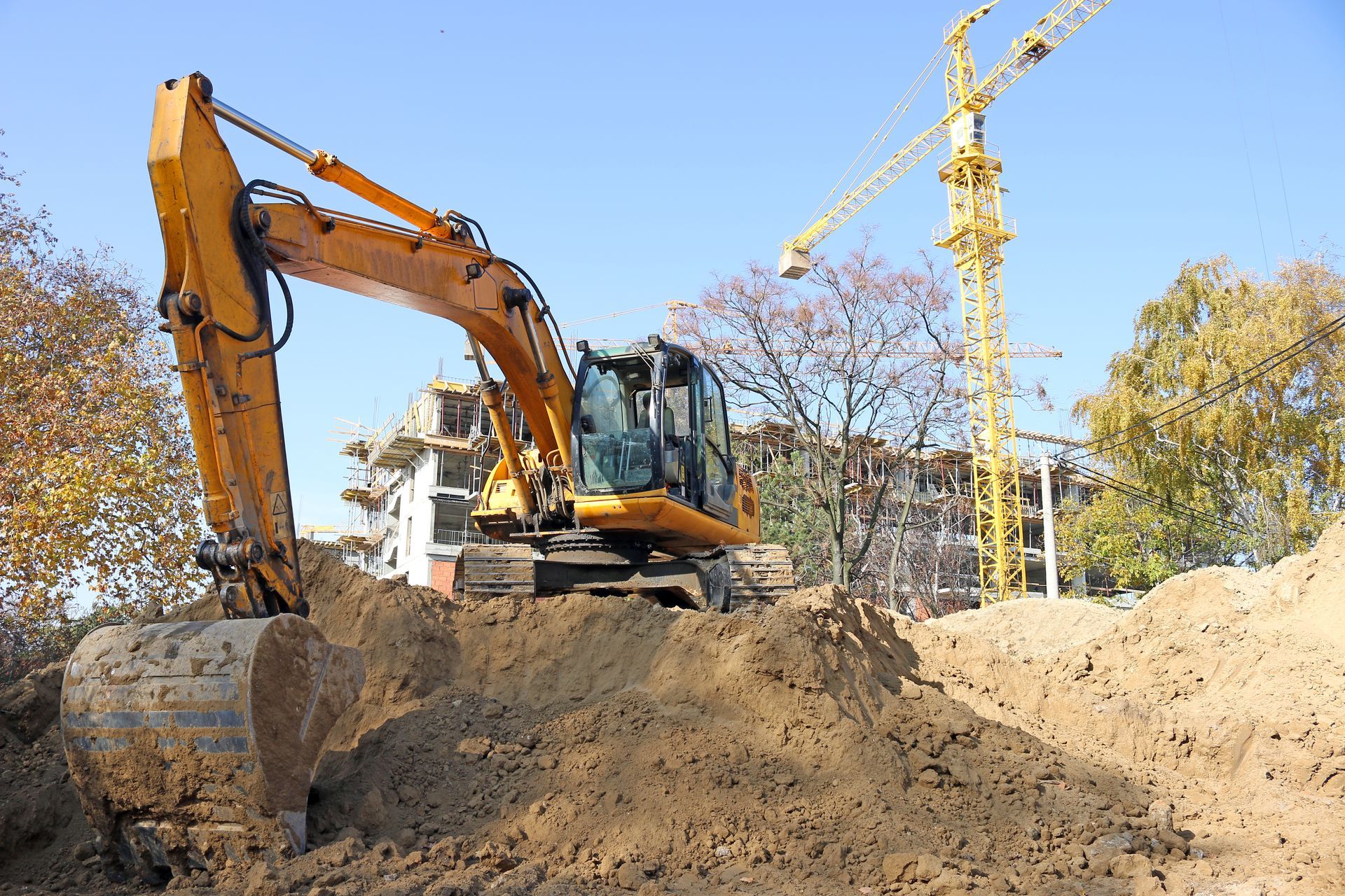 Yellow excavator digging a large dirt mound at a construction site with cranes in the background