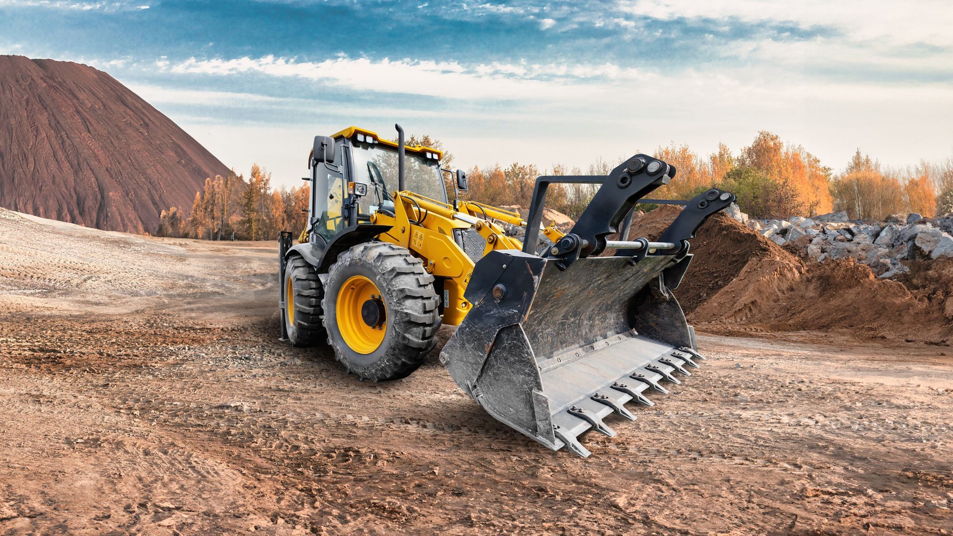 Yellow front loader with raised bucket on a dirt construction site near a gravel mound and autumn trees