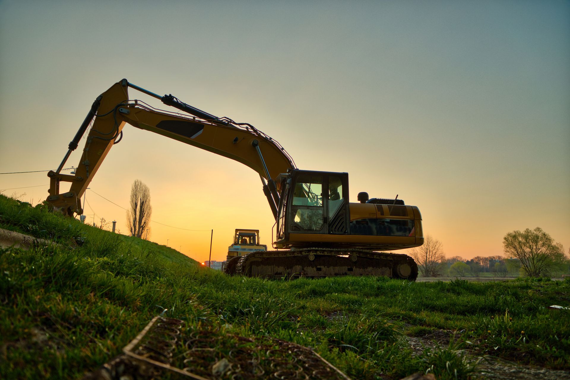 Excavator working on a grassy hillside at sunset, silhouetted against a warm sky.