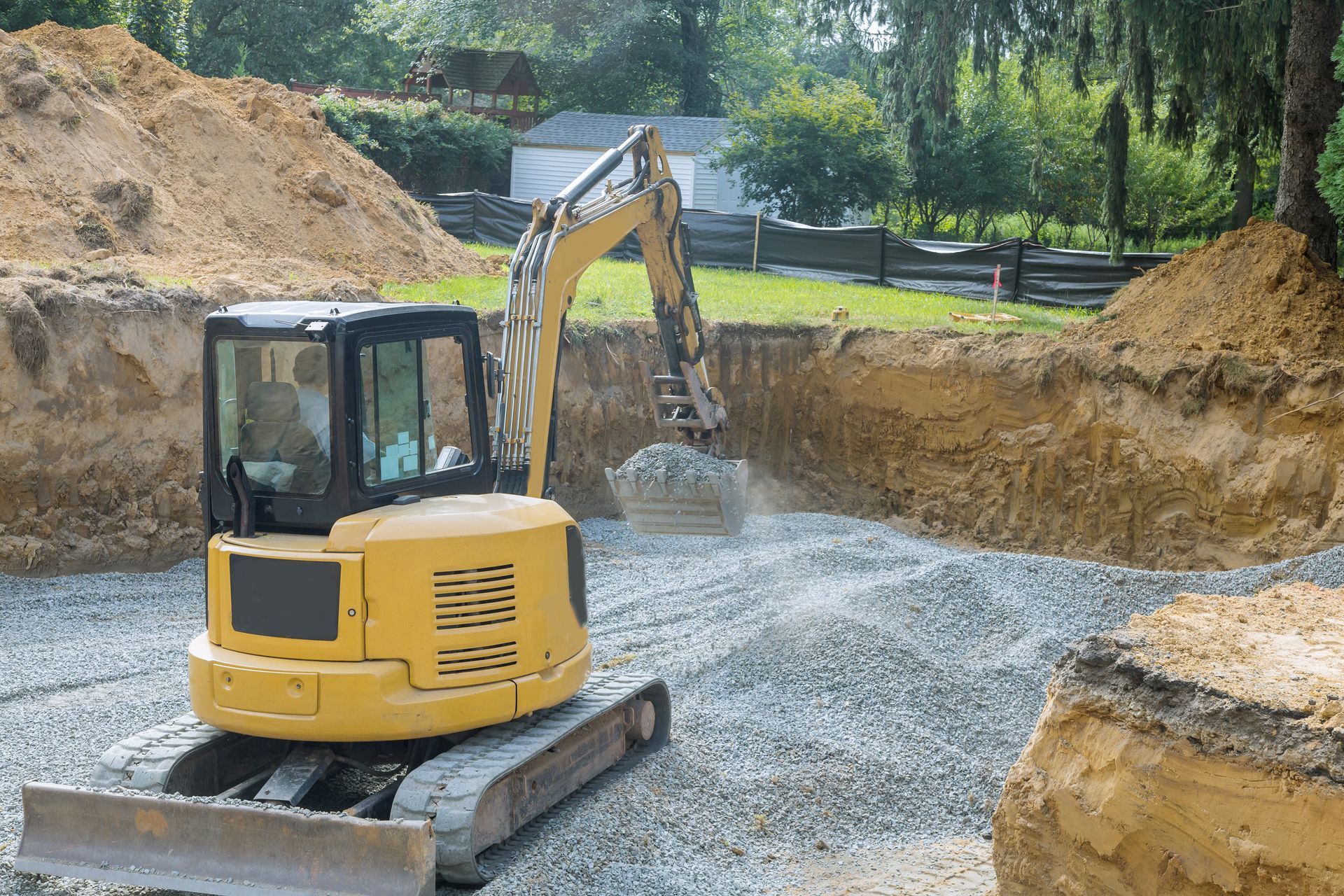 Yellow excavator digging gravel in a construction pit, surrounded by dirt mounds and trees.