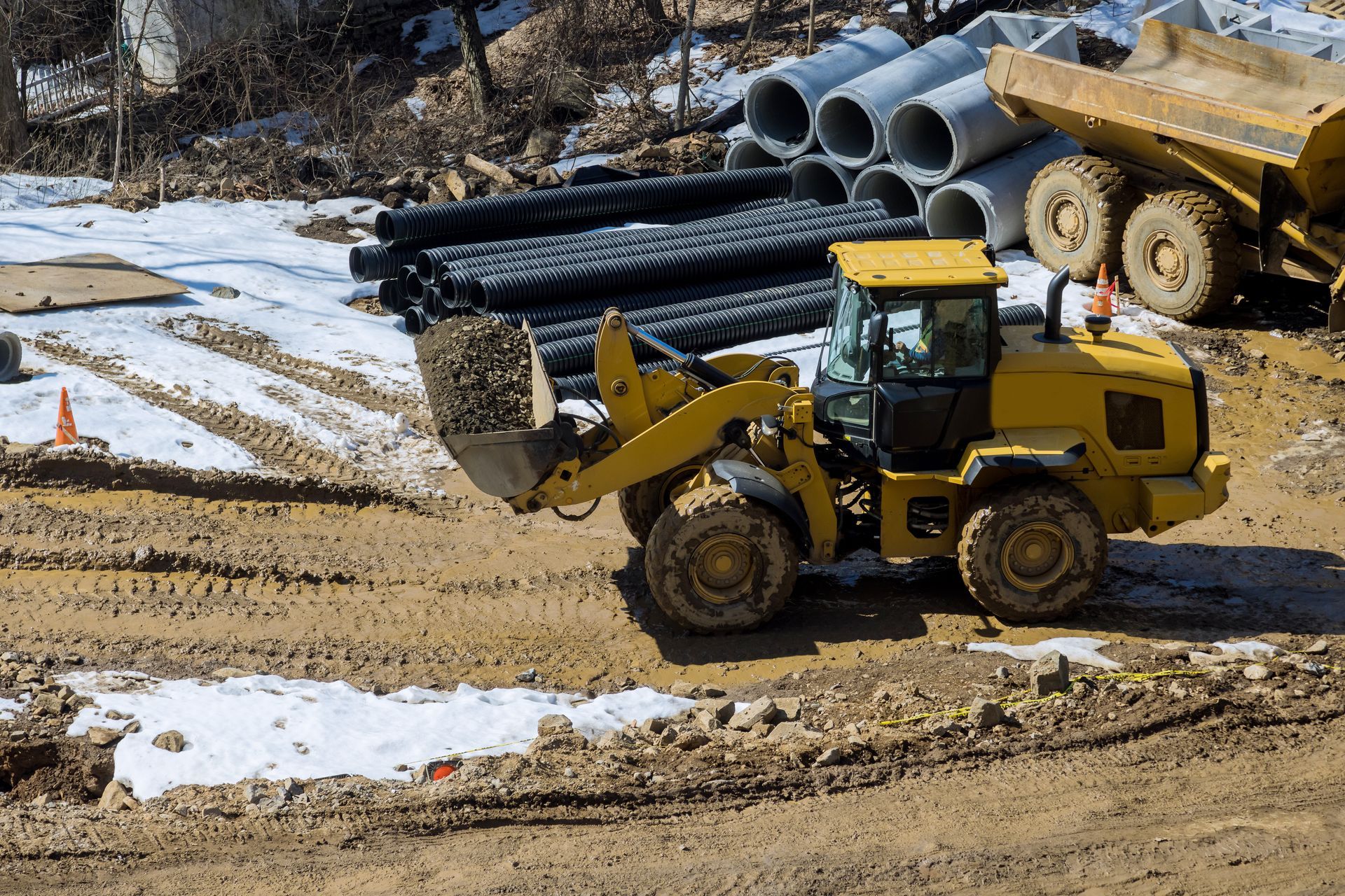 Yellow front loader moving dirt at a snowy construction site with pipes and trucks in the background
