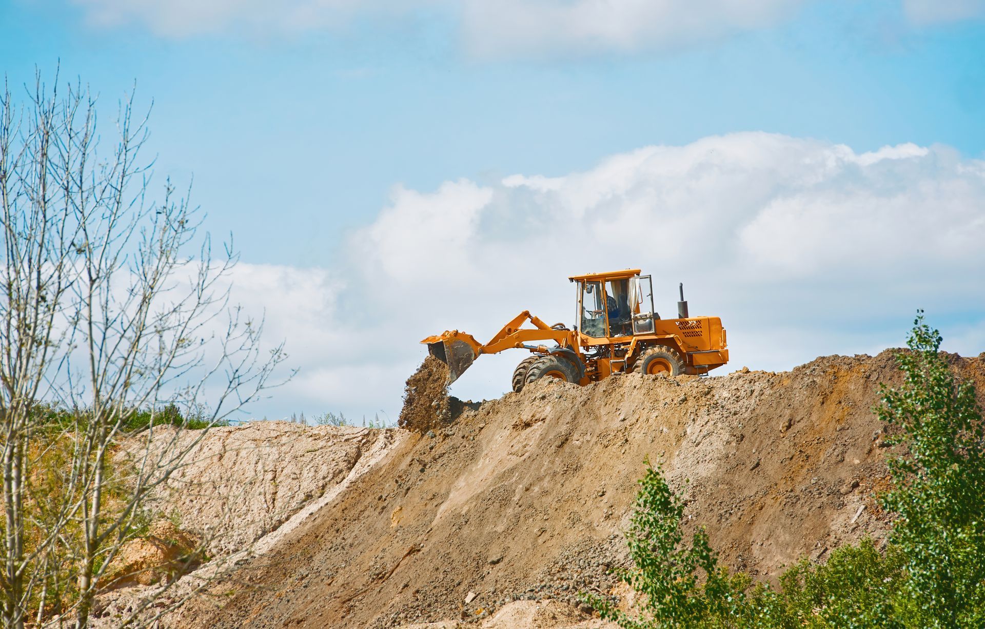 Yellow bulldozer digging soil on a dirt mound under a blue sky with clouds