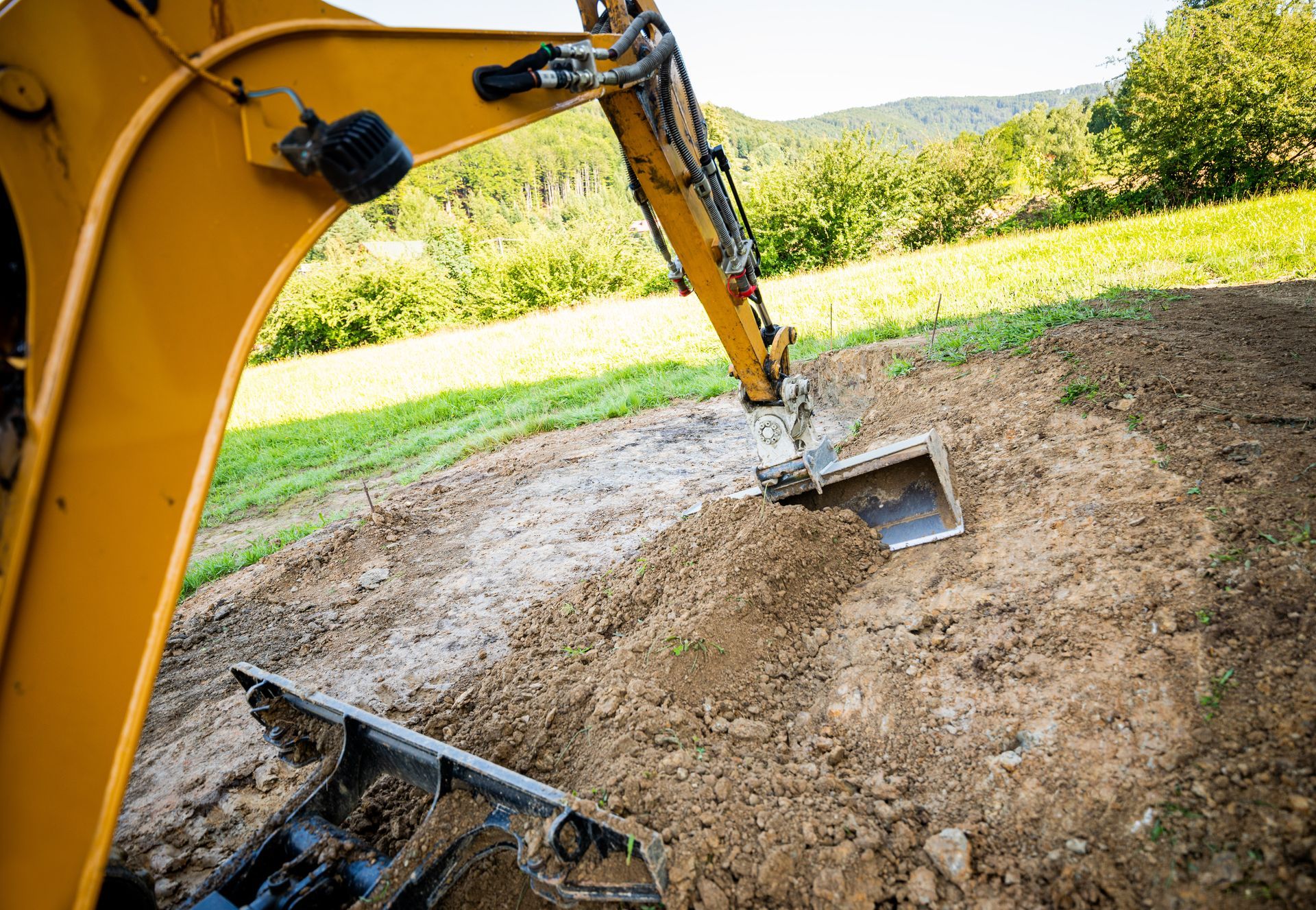 Yellow excavator digging a dirt mound on a grassy site, with a bucket in the foreground.
