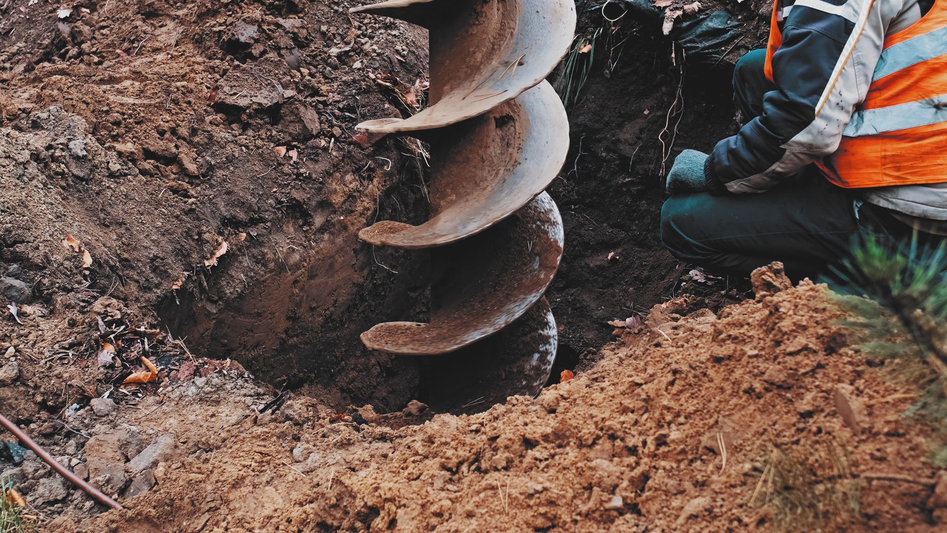 Large earth auger drilling soil beside a worker in orange safety gear at a construction site