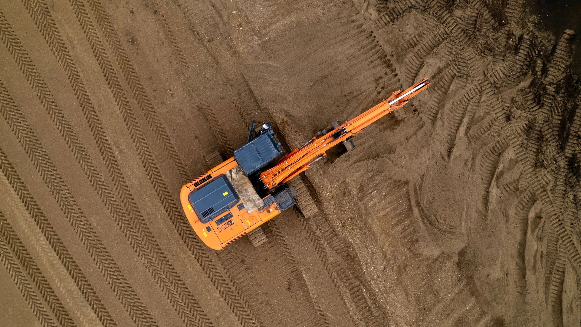 Orange excavator on brown dirt, viewed from above, with its arm extended across the ground.