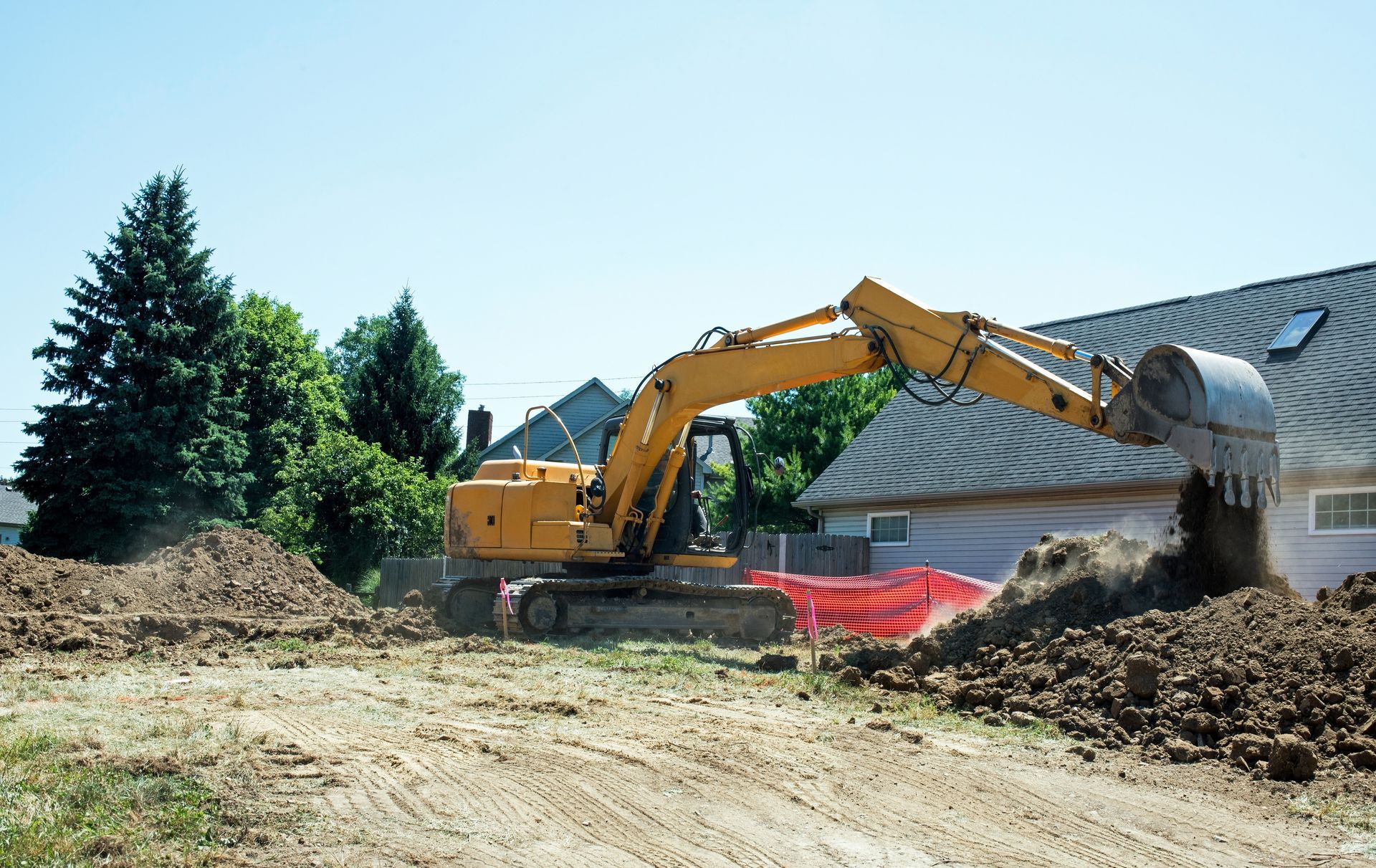 Yellow excavator moving dirt at a construction site beside a house and trees