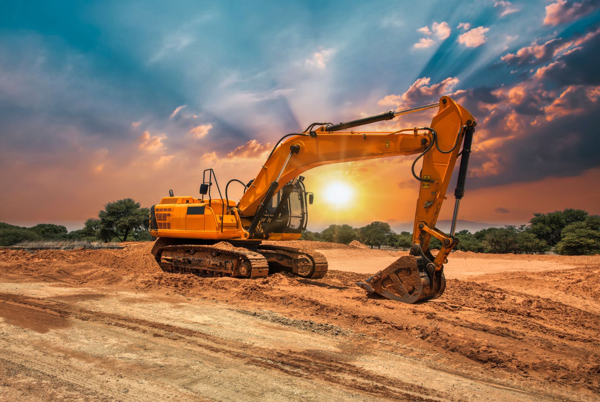 Yellow excavator working on a dirt site at sunset, with a warm orange sky.