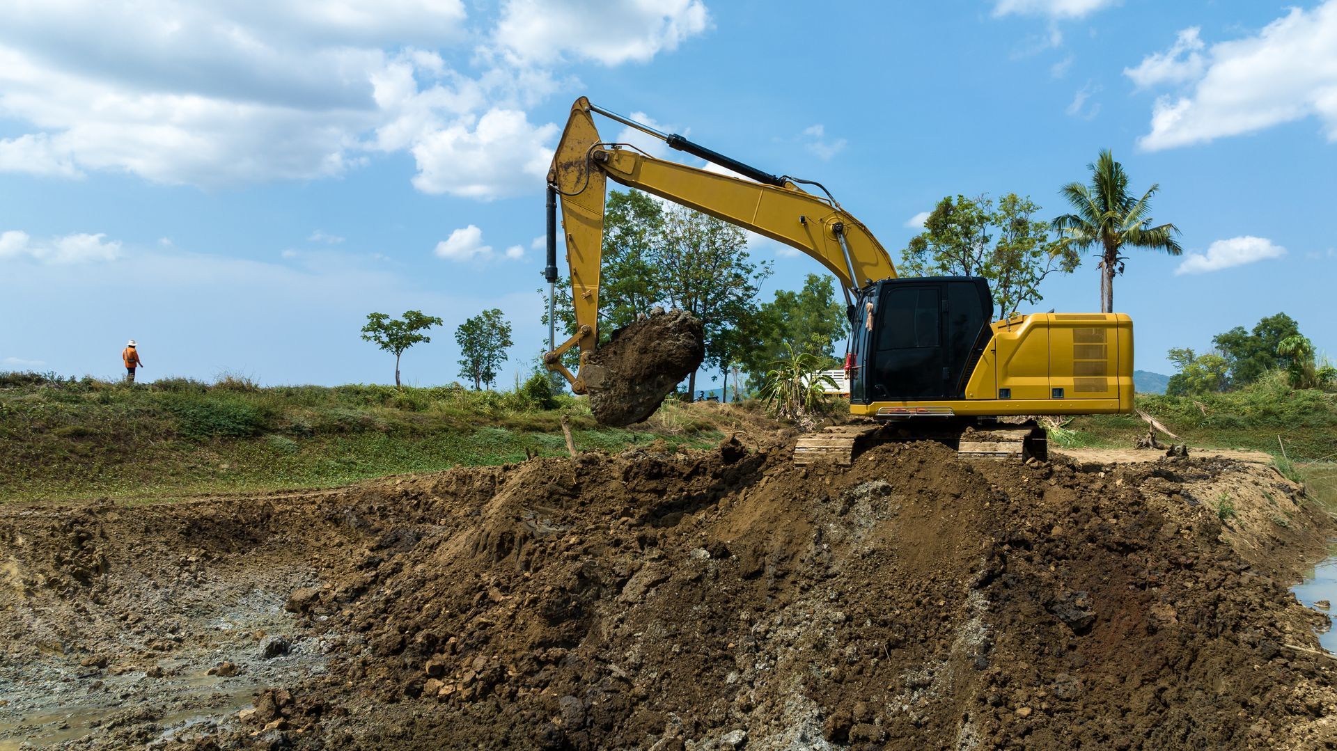 Yellow excavator digging a muddy hillside under a blue sky with scattered clouds