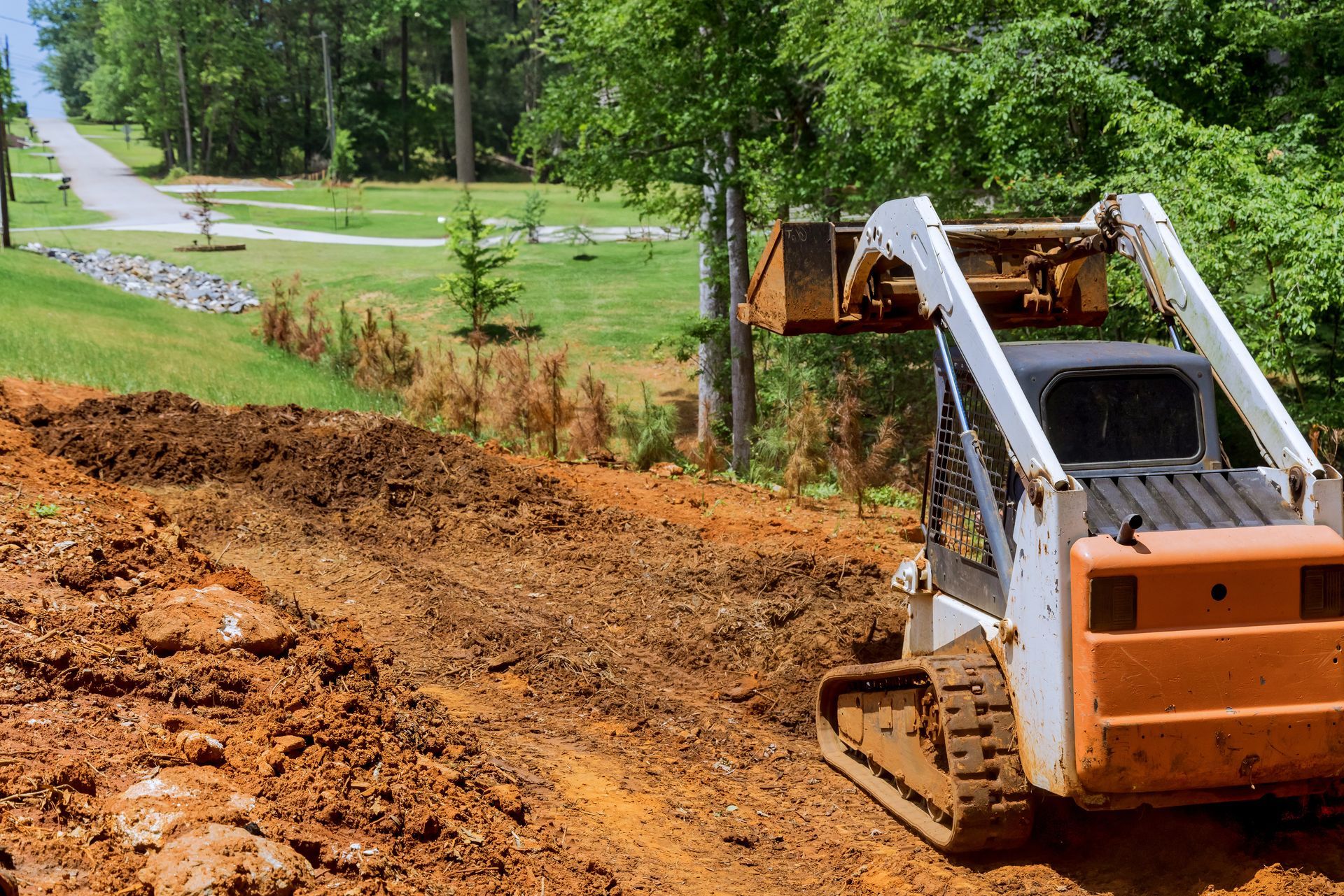 Orange skid steer on a muddy hillside near a grassy park path and trees