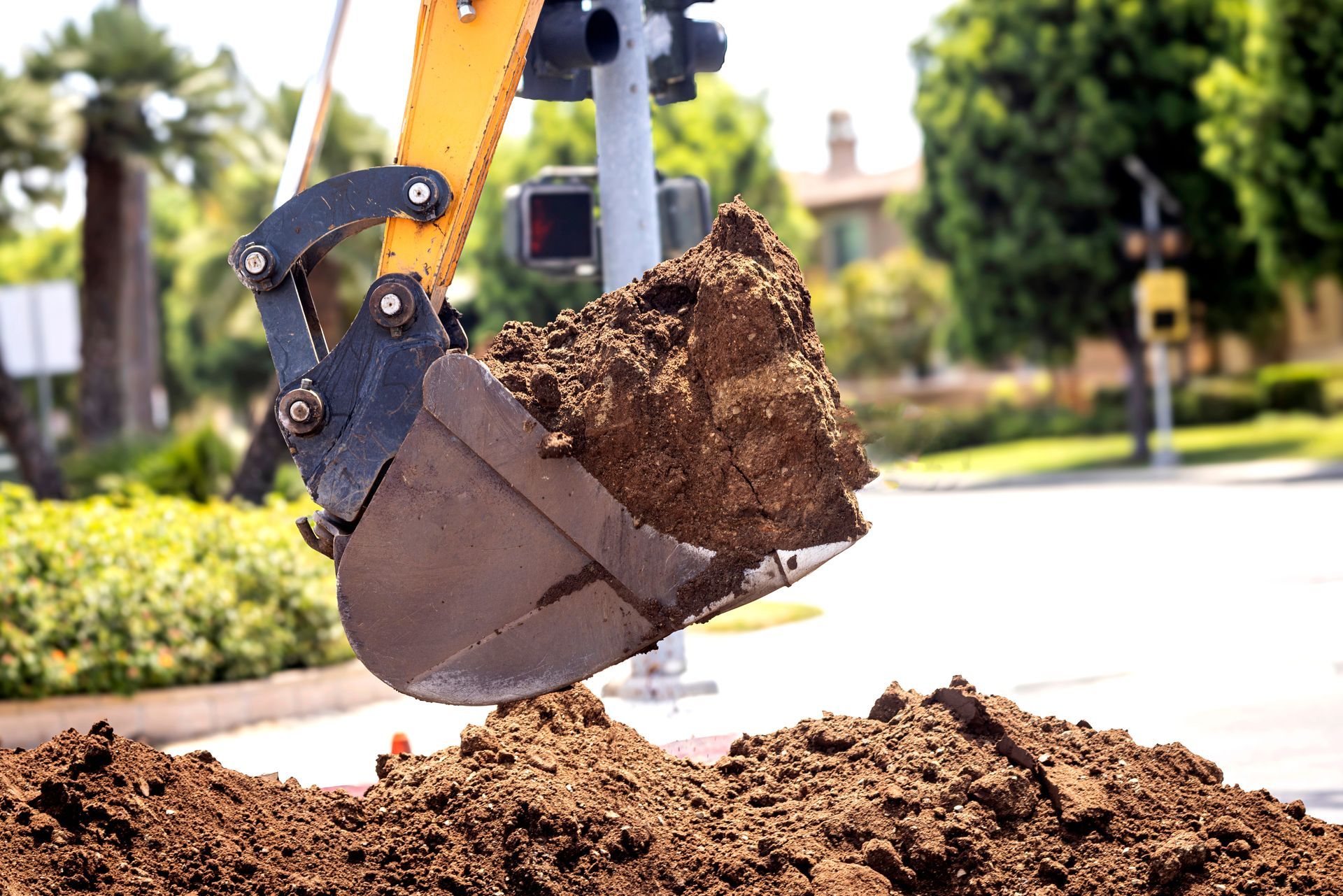 Backhoe bucket dumping dirt onto a pile beside a road in bright daylight
