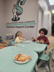 Two little girls are sitting at tables eating pizza.