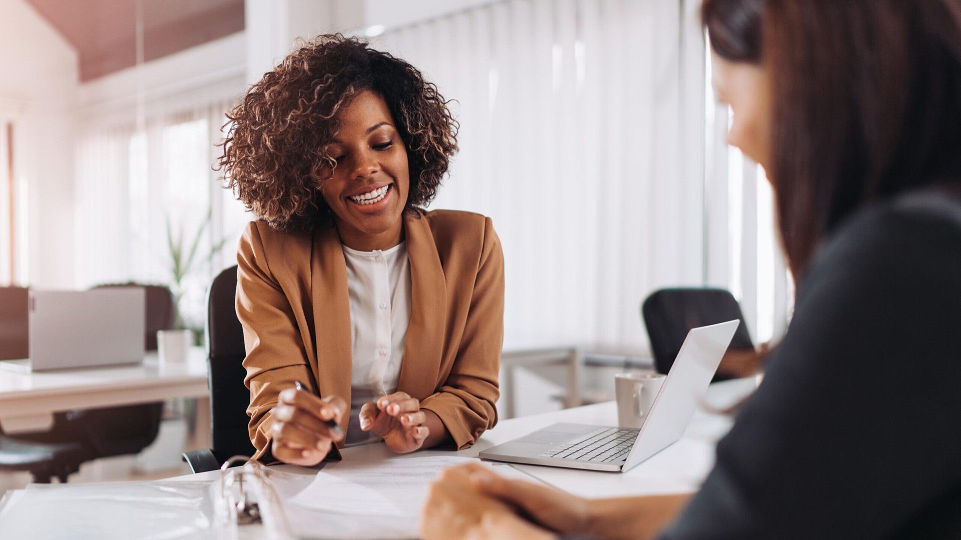 Une femme en blazer beige sourit et fait des gestes tout en discutant d'un document avec une personne assise à un bureau.