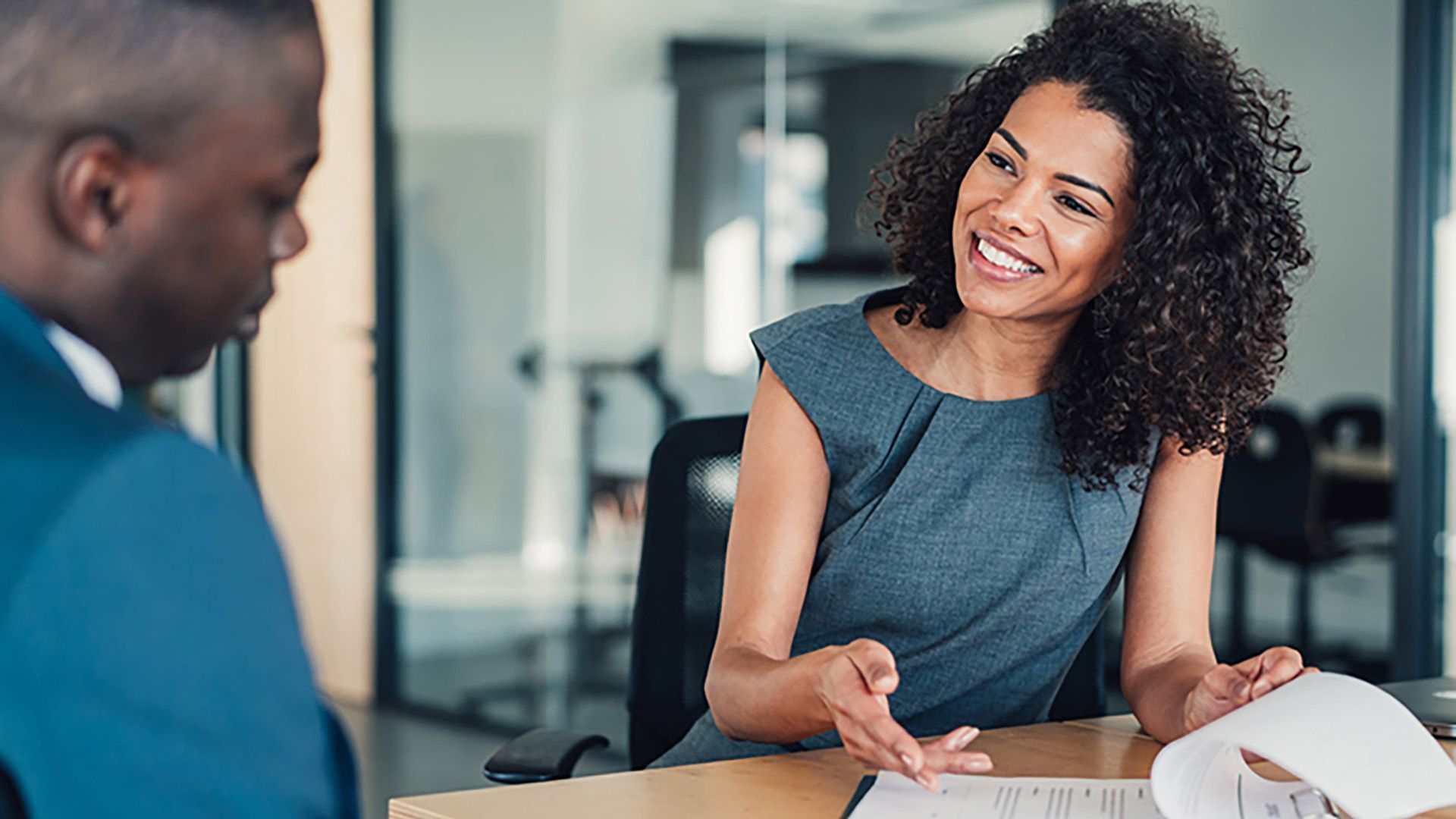 Femme souriante, faisant des gestes, examinant des documents avec une personne dans un environnement de bureau moderne.