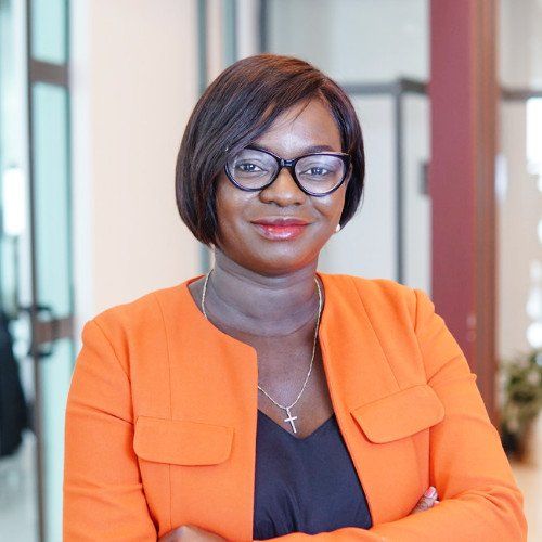 Une femme portant des lunettes et une veste orange est debout avec les bras croisés.