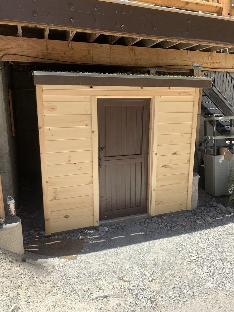 Small light-wood shed with a brown door under a deck, beside gravel and utility units.
