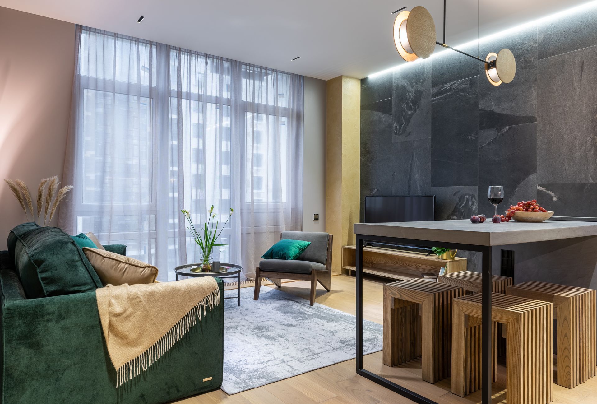 Modern living room with green sofa, large window, glass dining table, and wooden stools