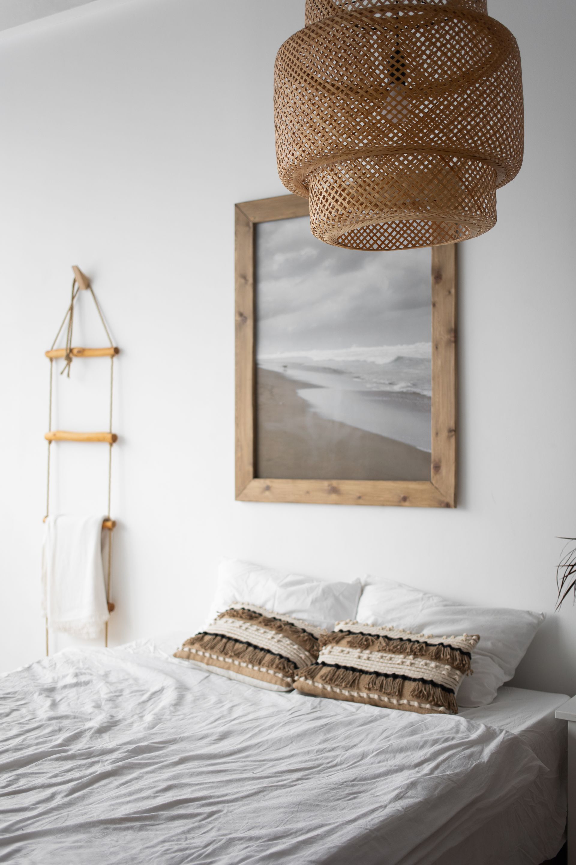 Cozy neutral bedroom with white bedding, woven pendant light, and framed beach landscape above the bed