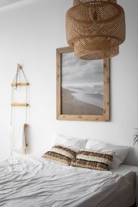 Cozy neutral bedroom with white bedding, woven pendant light, and framed beach landscape above the bed