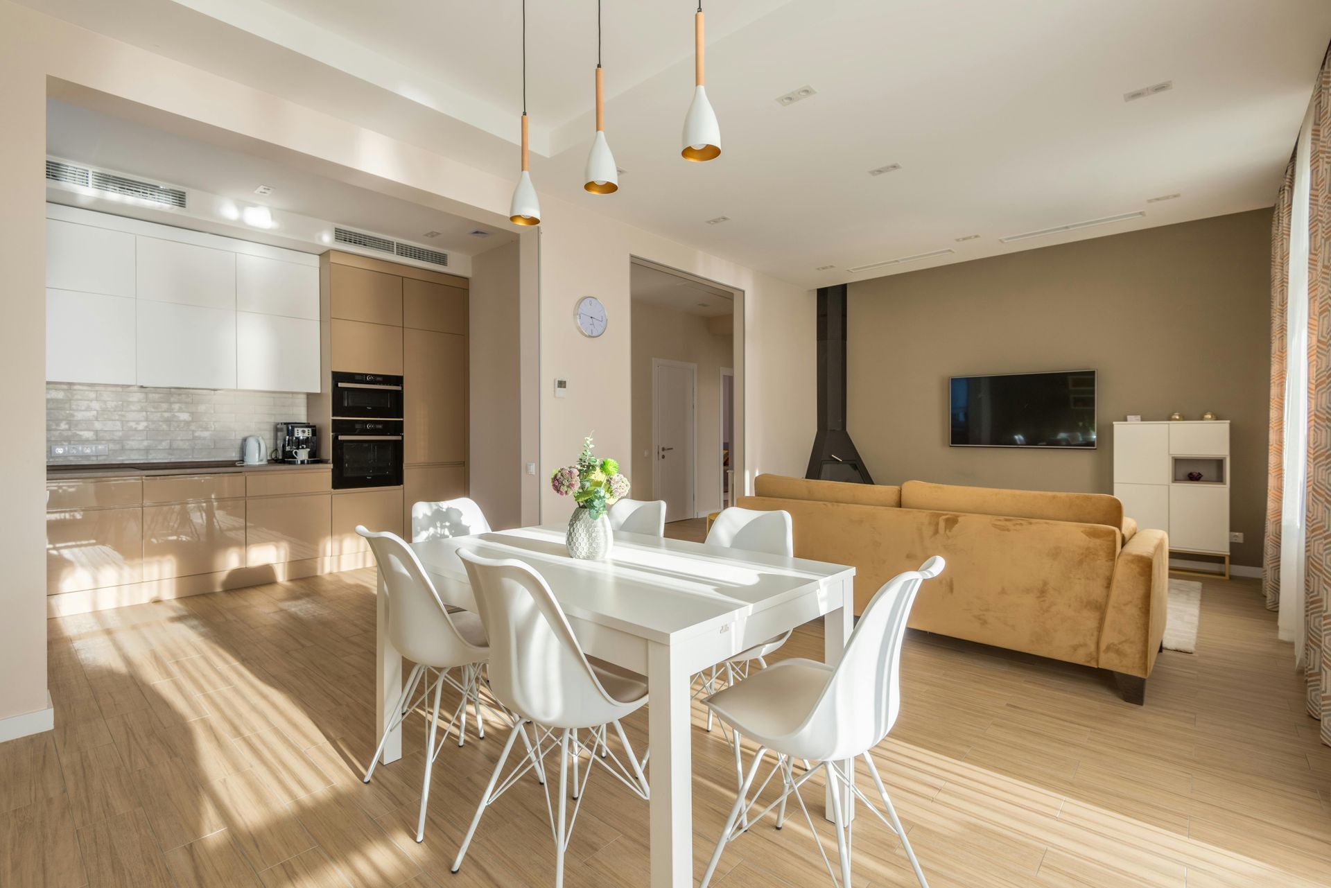 Bright open-plan dining and living room with white table, tan sofa, wall-mounted TV, and kitchen.