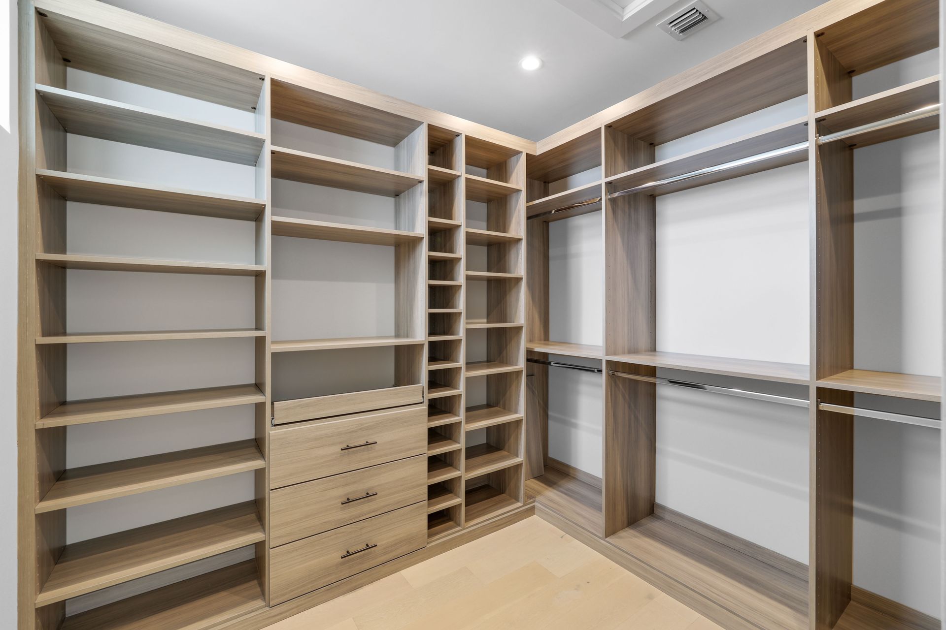 Spacious walk-in closet with empty light wood shelving, drawers, and hanging rods in a neutral room