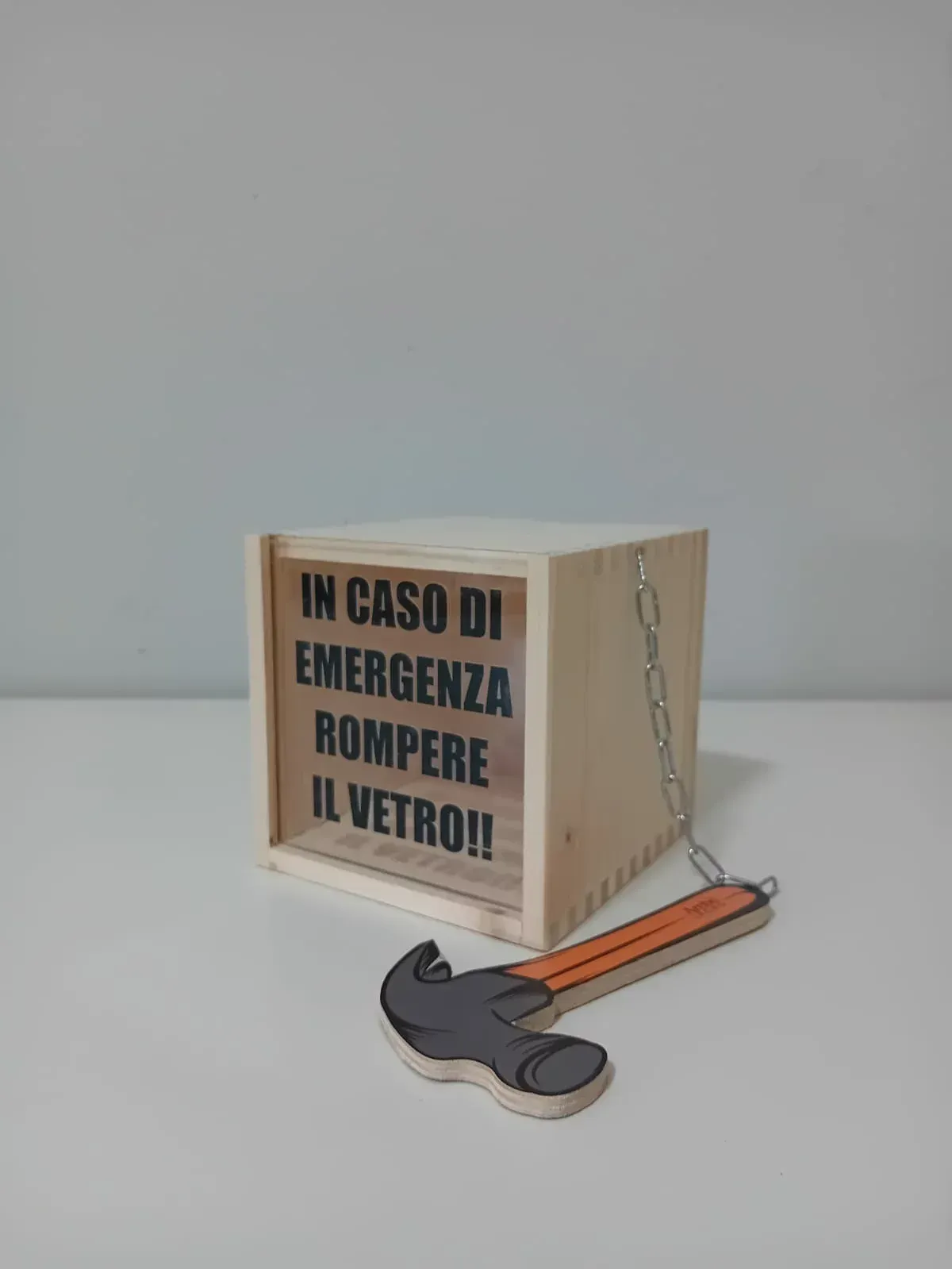 Cassetta di emergenza in legno con etichetta in italiano e un martello accanto, su sfondo grigio.