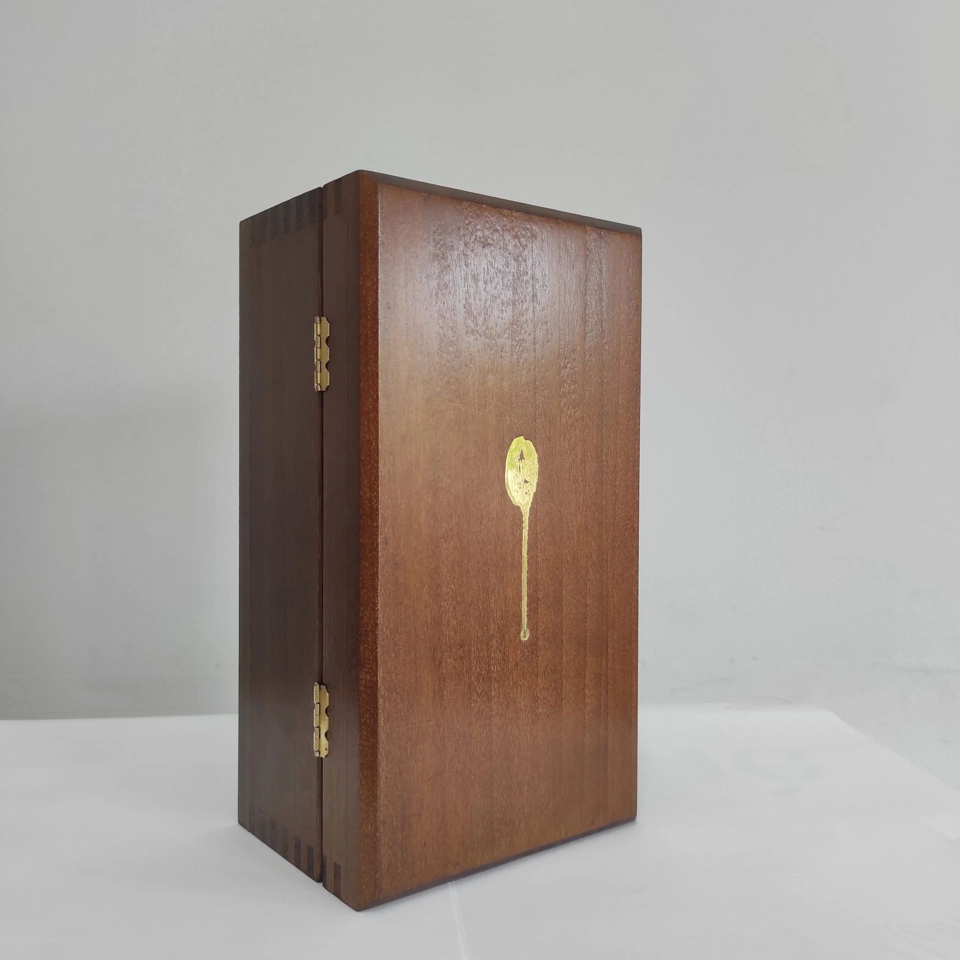 Scatola in legno a forma di libro con cerniere in ottone e un piccolo emblema dorato sul davanti, poggiante su una superficie leggera.