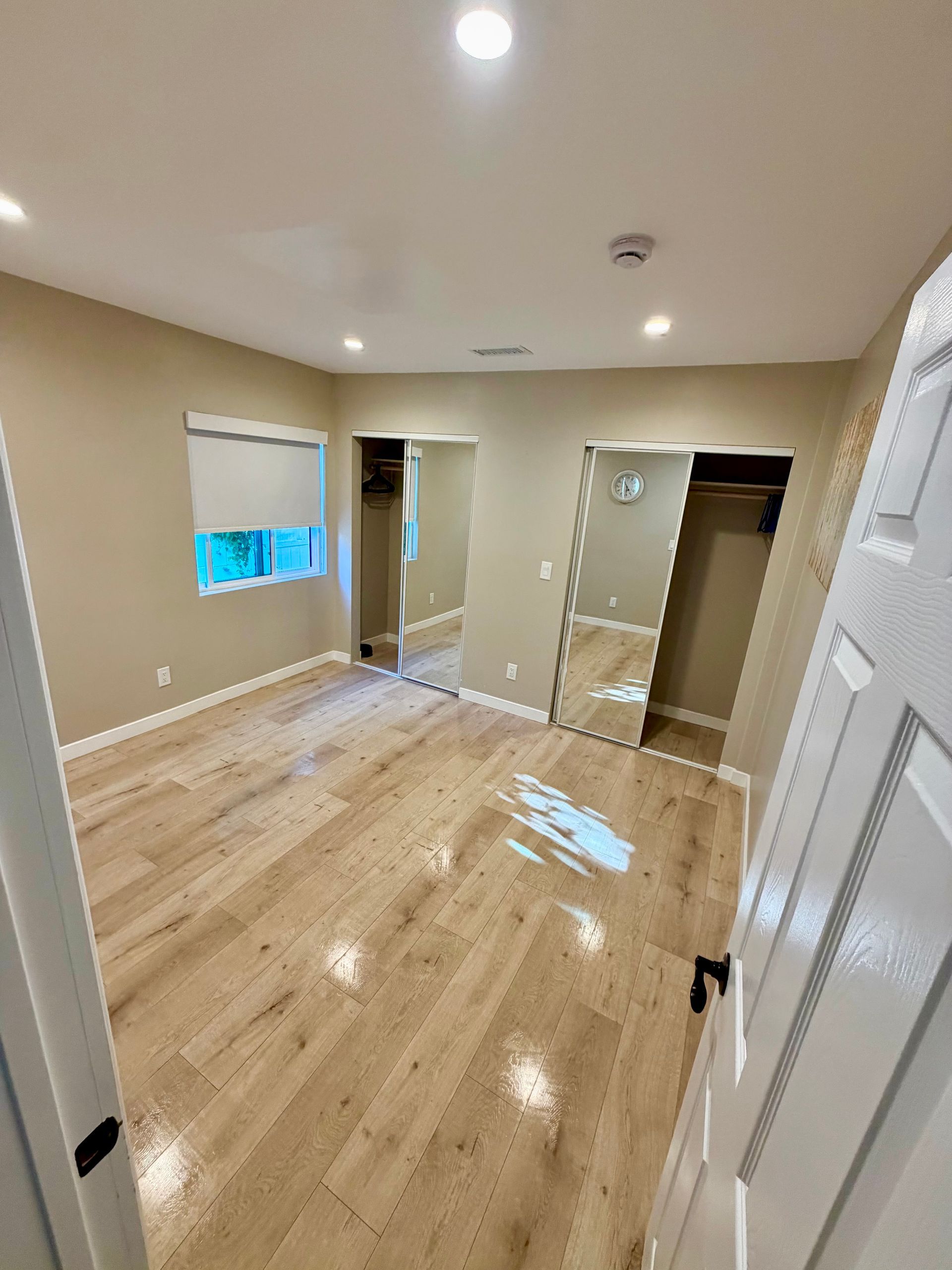 Empty beige room with hardwood floors, recessed lights, a window, and an open doorway to a closet or bathroom.