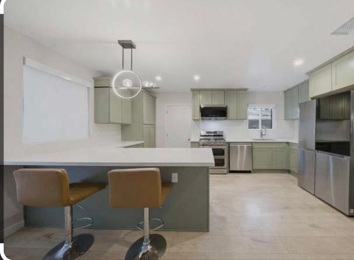 Modern kitchen with island, two tan stools, pale green cabinets, stainless steel appliances, and bright lighting