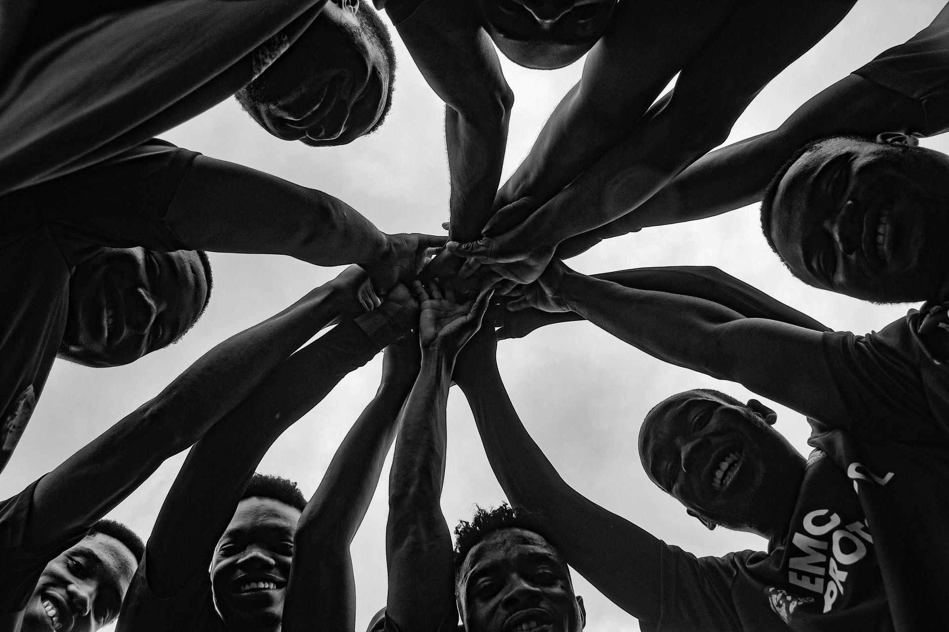 Group of people standing in a circle, arms extended inward against a bright sky, viewed from below