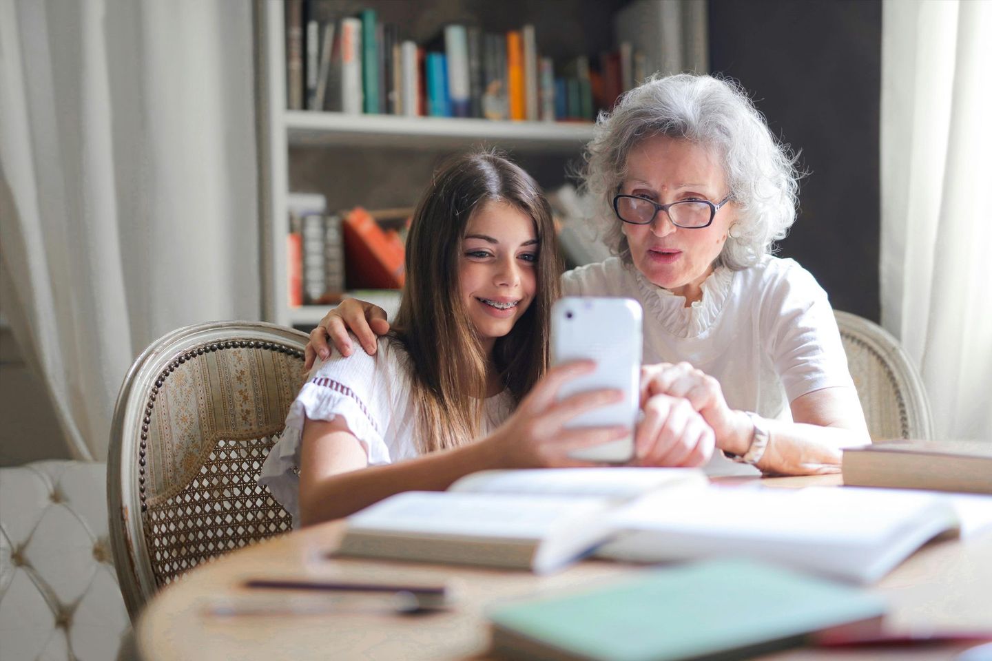 Two people sitting at a table together, looking at a smartphone screen with focused expressions. Books are on the table.