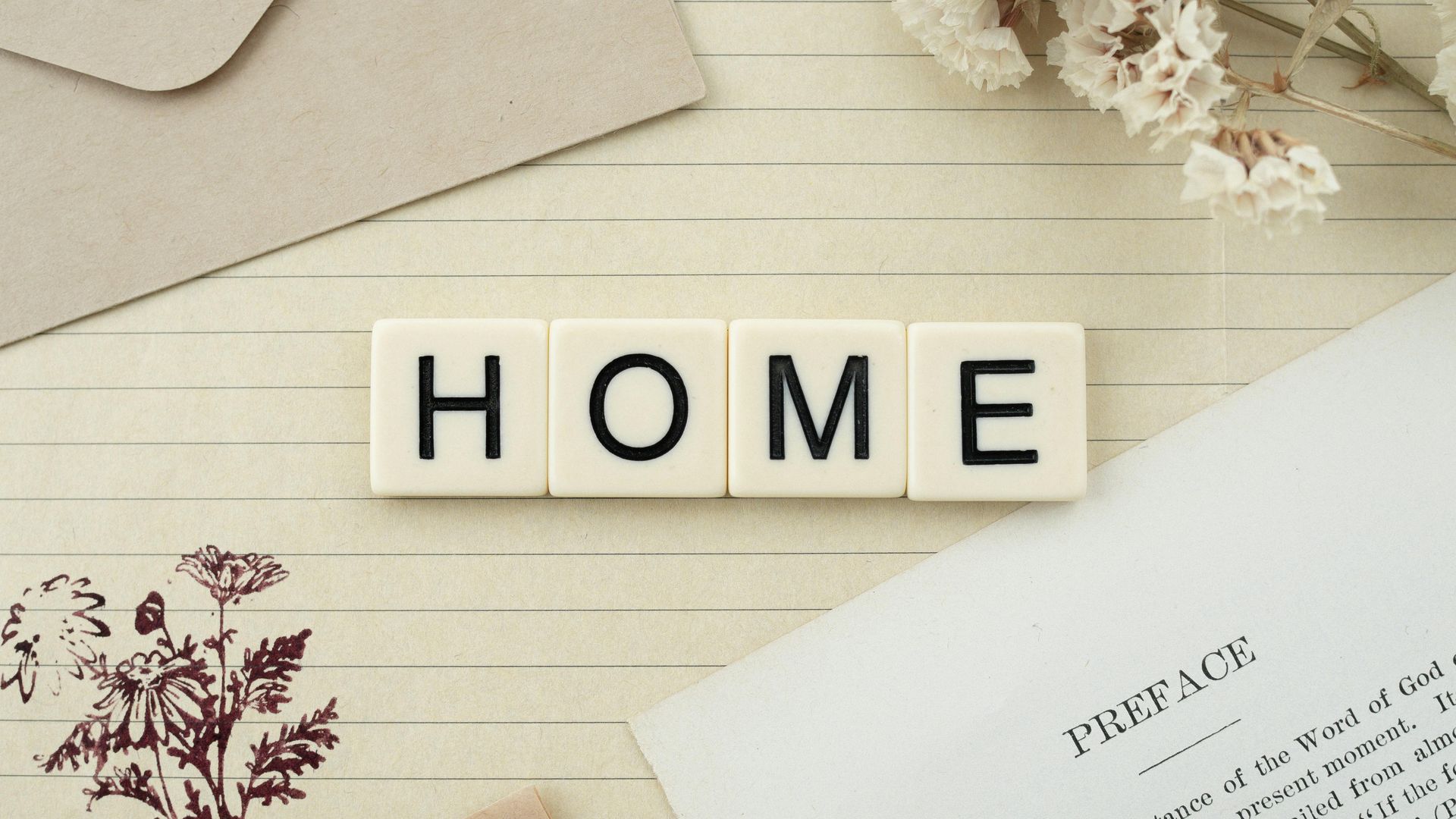 Cream tiles spelling “HOME” on a beige tabletop with a folded paper and dried flowers.
