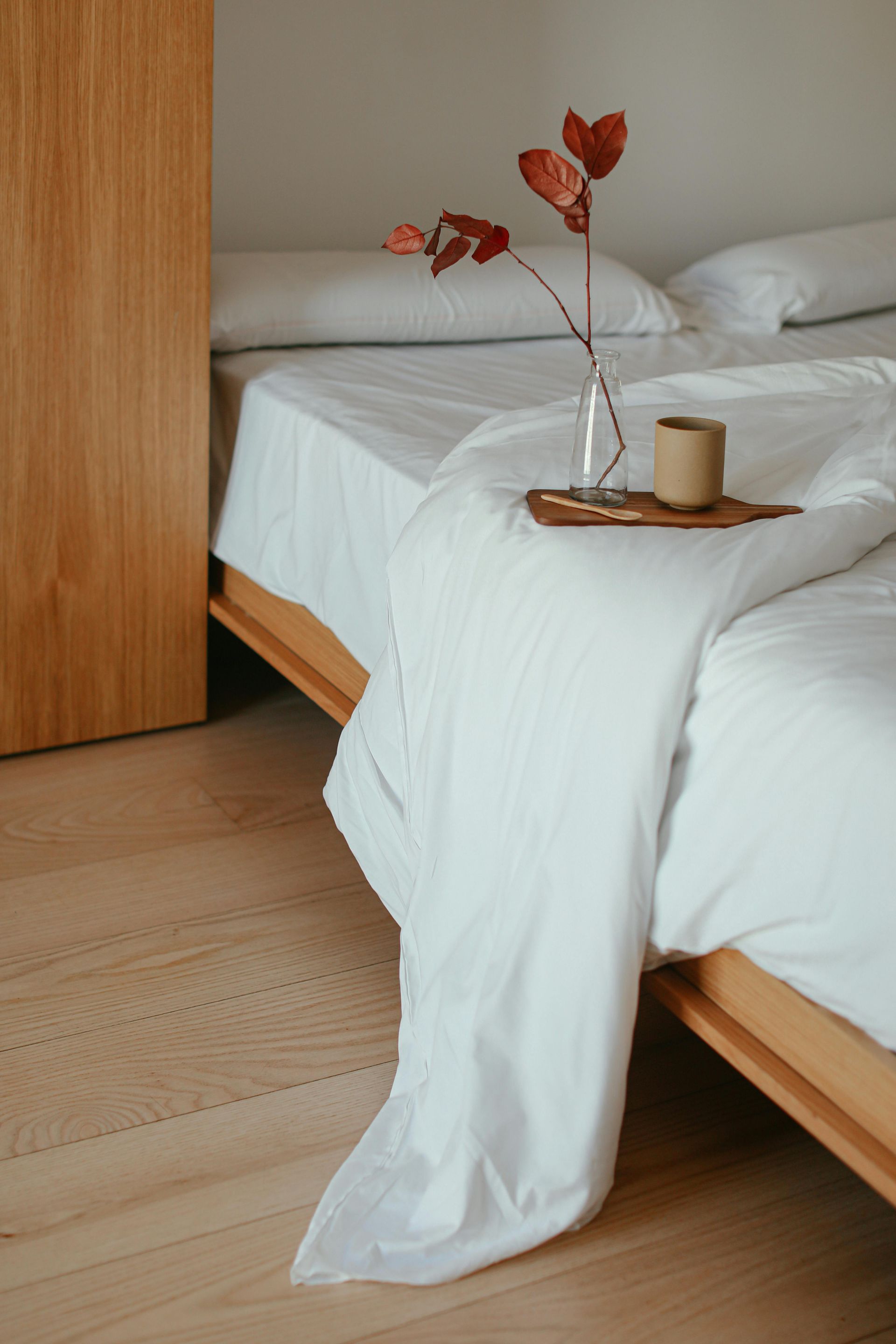 Minimalist bedroom with a white bed, wooden floor, and a tray holding a vase and candle on the draped duvet