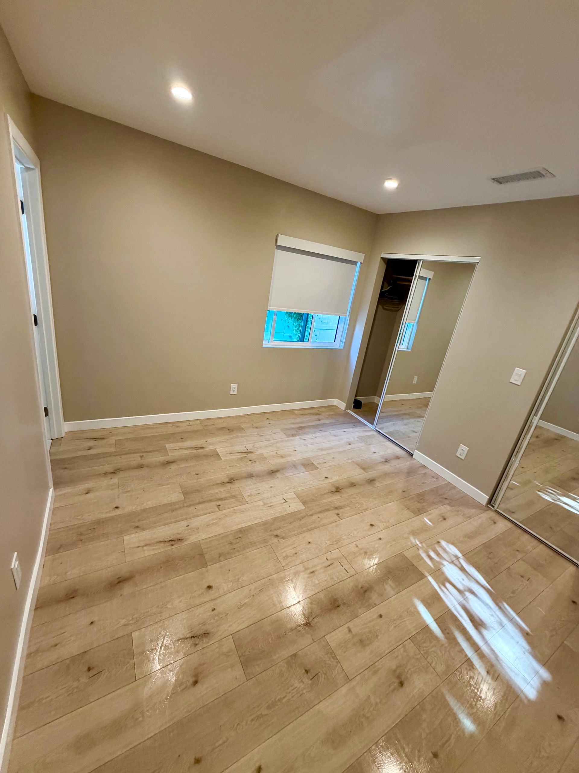 Empty beige bedroom with light wood floors, a window, recessed lights, and an open doorway to a closet.