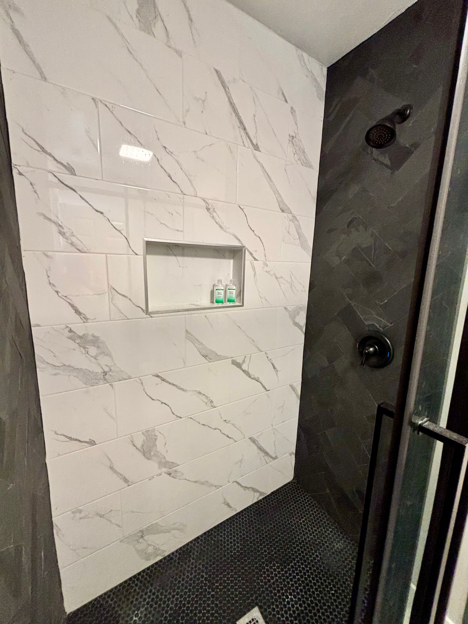Modern shower stall with marble wall tiles, black textured floor, and a recessed shelf holding green bottles