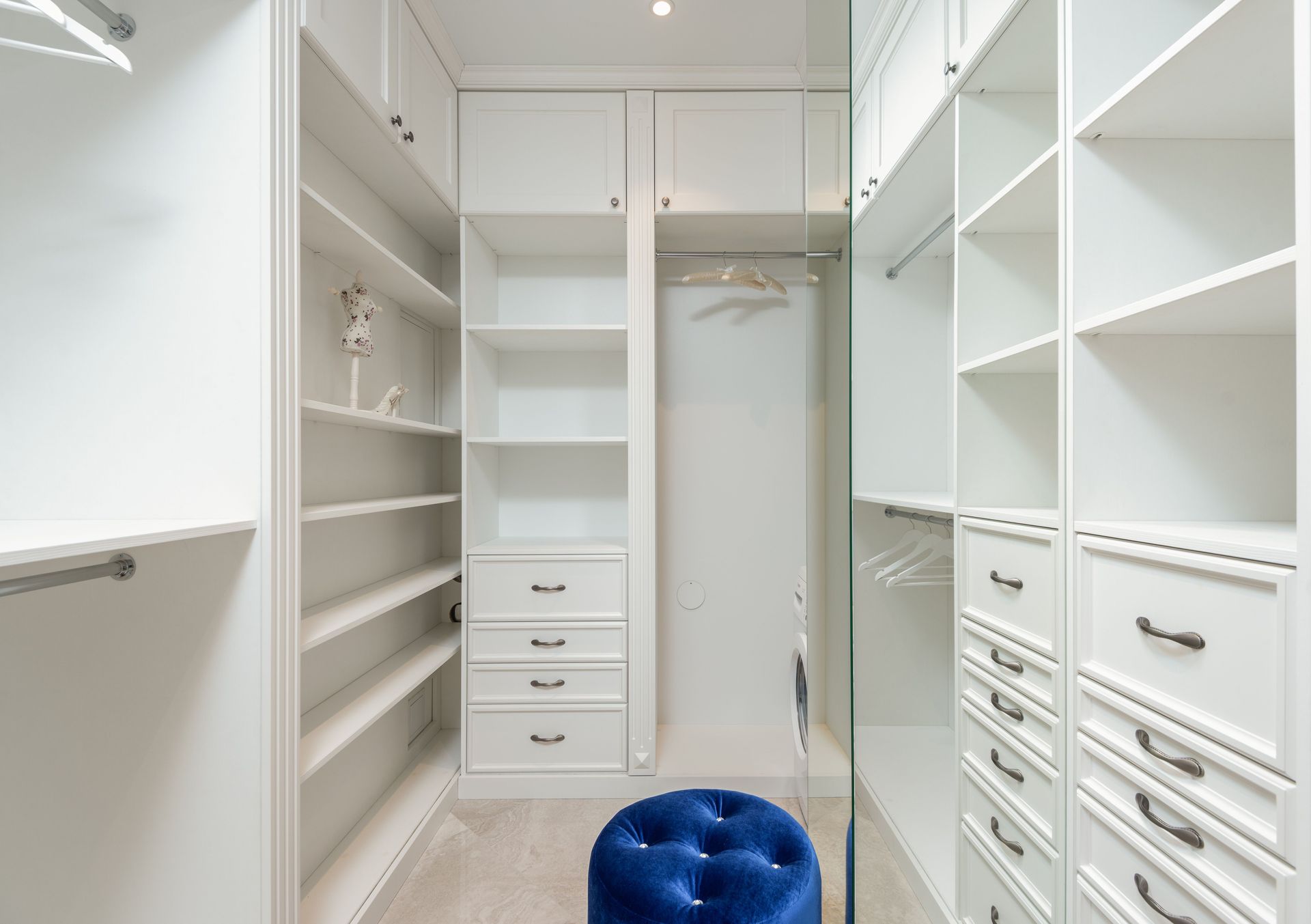 Bright white walk-in closet with built-in shelves and drawers, and a blue ottoman in the center.