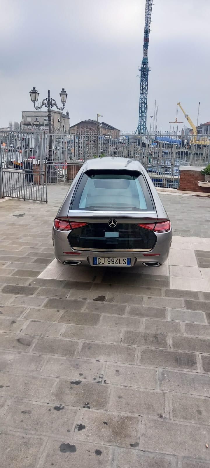 Vista posteriore di un carro funebre argentato parcheggiato su una strada acciottolata di fronte a un paesaggio urbano sotto un cielo nuvoloso.