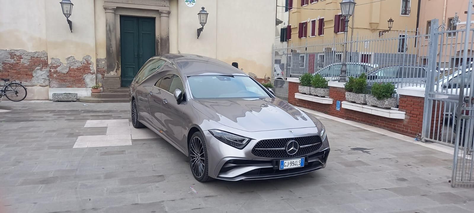 Una station wagon Mercedes-Benz grigia parcheggiata in una piazza acciottolata di fronte a un edificio con una porta verde.