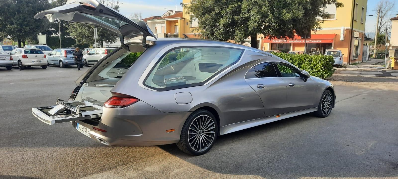 Carro funebre grigio con portellone posteriore aperto, parcheggiato su una strada asfaltata, di fronte a degli edifici, di giorno.