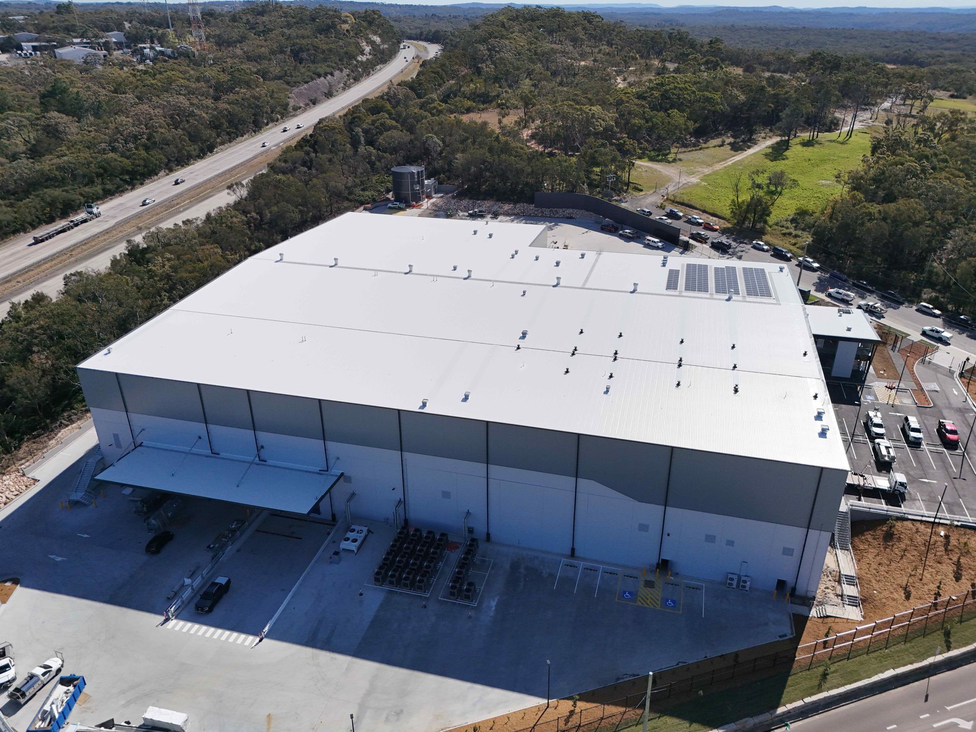 Large industrial warehouse with gray and white exterior, near a highway and park.