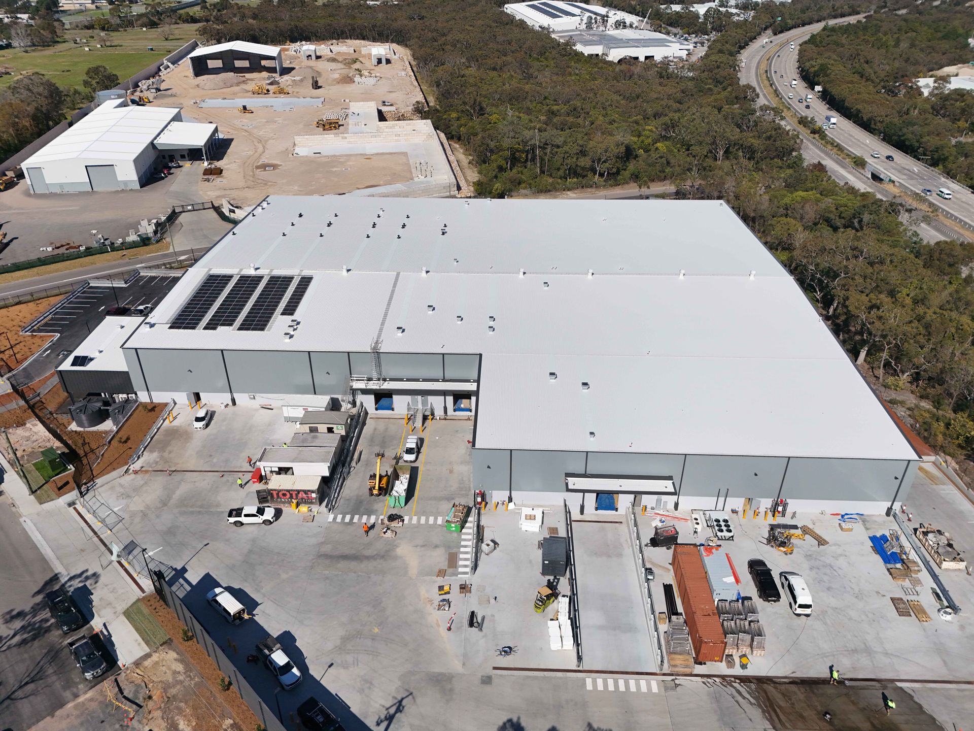 Aerial view of a large warehouse with solar panels and loading docks. Construction site beside it.