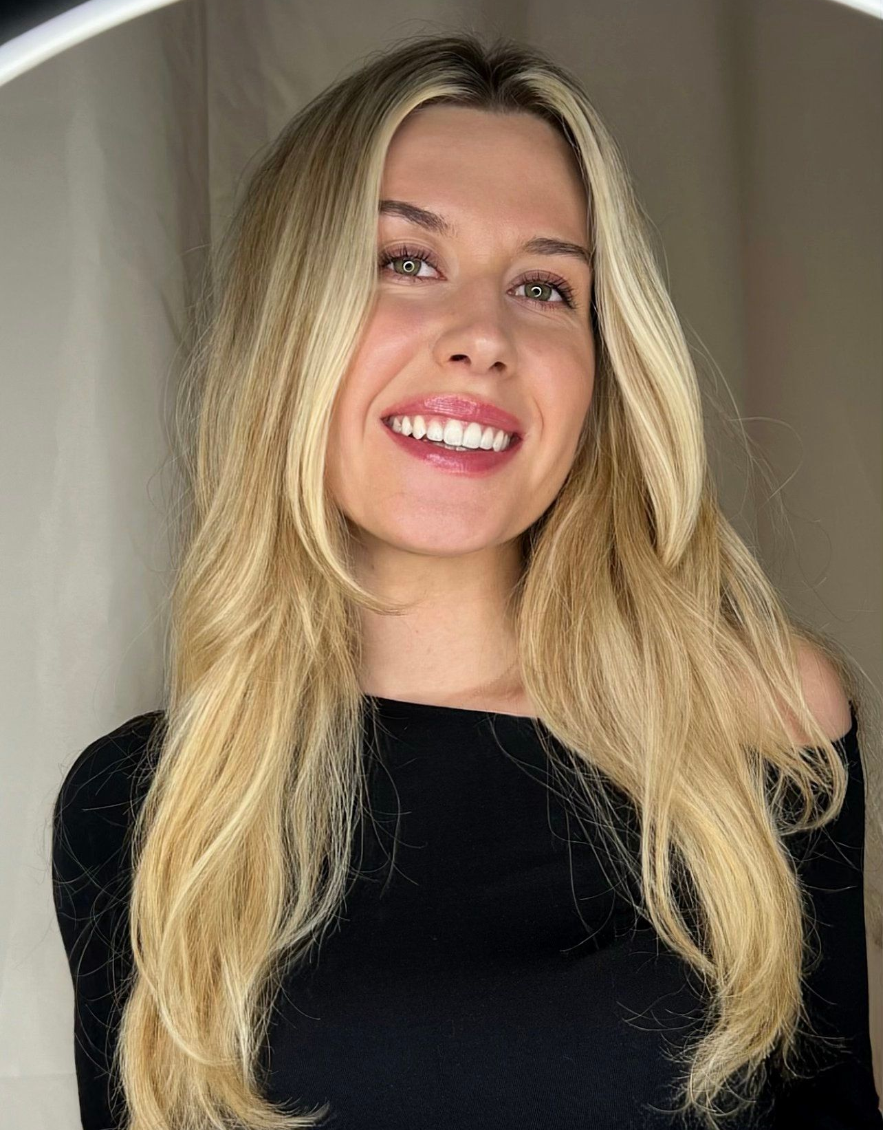 Lächelnde blonde Frau, trägt ein schwarzes Oberteil. Langes, welliges Haar, sanftes Licht.